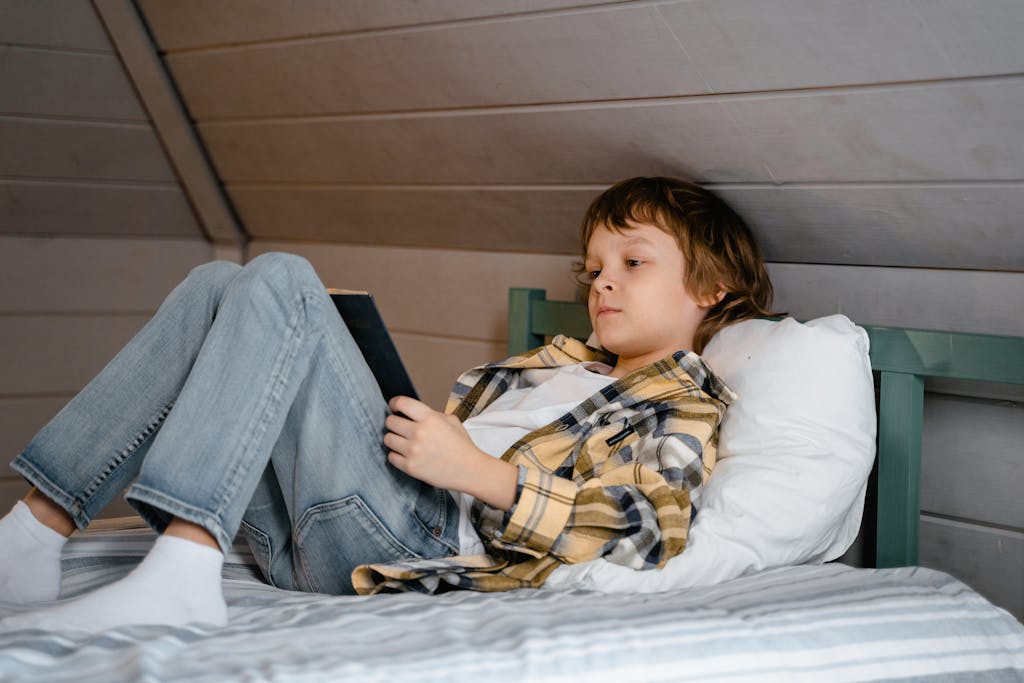 A young boy is lying on a bed reading a book in a cozy, wooden-themed bedroom.